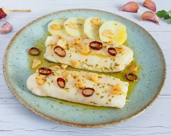 Dos filetes de merluza a la bilbaina, con patatas, ajo laminado y guindilla, en un plato azul.