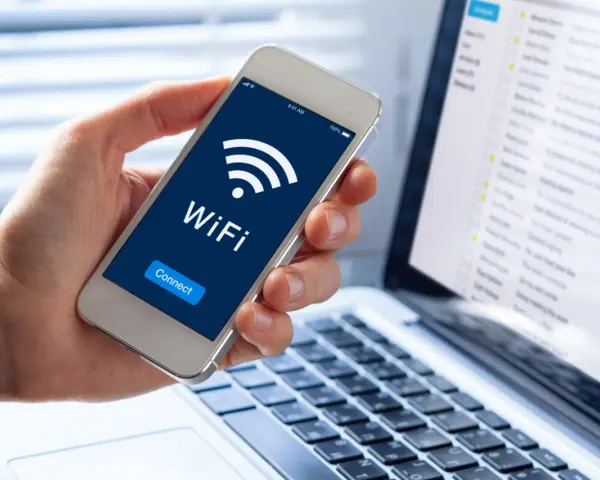 Dłoń trzyma smartfon z ikoną WiFi i napisem "Connect". Obok widać laptopa z listą e-maili, sugerującą pracę zdalną i dostęp do sieci, być może przez **ssid** domowy.