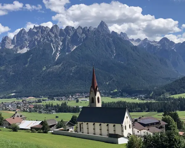 Chiesa con campanile rosso in primo piano, sullo sfondo le maestose vette delle Dolomiti e i **paesi più belli della Val Pusteria**.