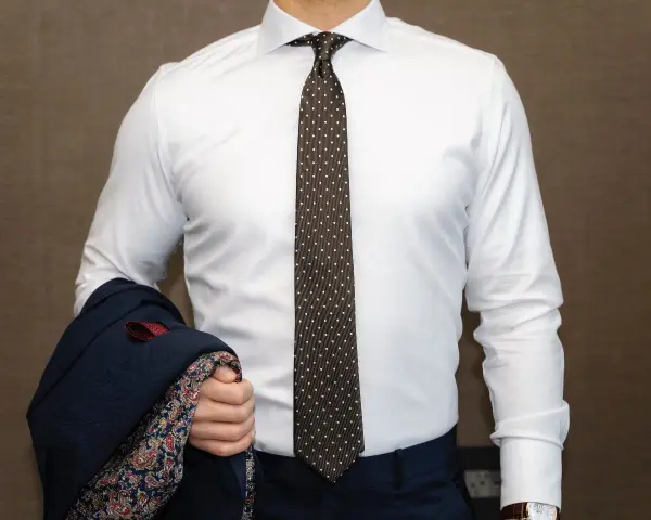 Elegancki mężczyzna w białej koszuli i modnym krawacie męskim w kropki, trzymający granatową marynarkę z wzorzystą podszewką.