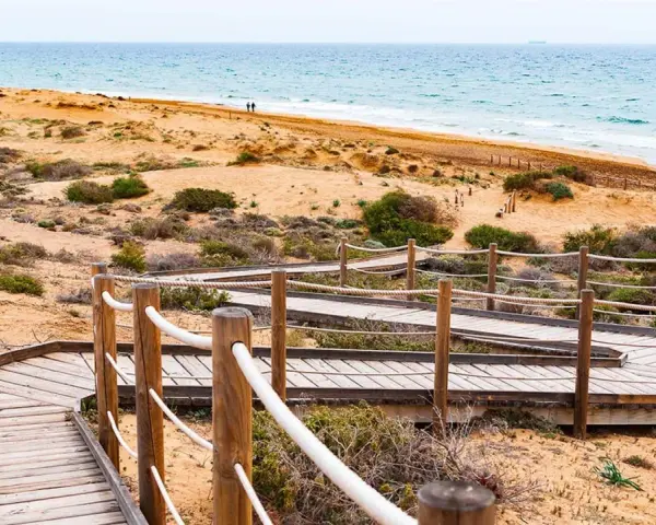 Pasarela de madera serpentea por la duna, invitando a explorar la ruta Calblanque hacia el mar azul.
