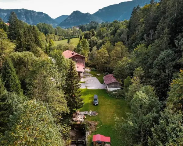 Idyllisches **Camping in den Bergen**: Ein Wohnmobil steht auf einer grünen Wiese neben einem Bach, umgeben von dichten Wäldern und traditionellen Holzhäusern.