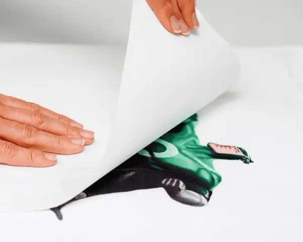 Manos aplicando una hoja para imprimir transfer sobre una camiseta blanca con un diseño de superhéroe verde.