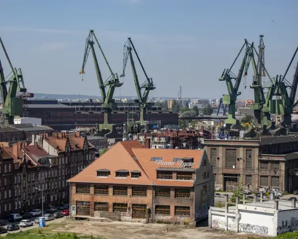 Widok na stocznię w Gdańsku z charakterystycznymi dźwigami. W oddali budynek z napisem "Nowe Syrie", nawiązujący do muzeum sztuki współczesnej Gdańsk.