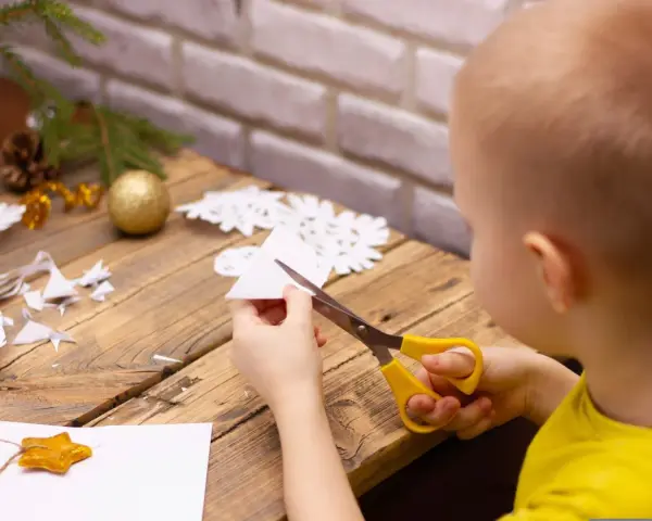 Małe rączki wycinają śnieżynkę z papieru. Na stole leżą gotowe ozdoby i gałązka choinki.