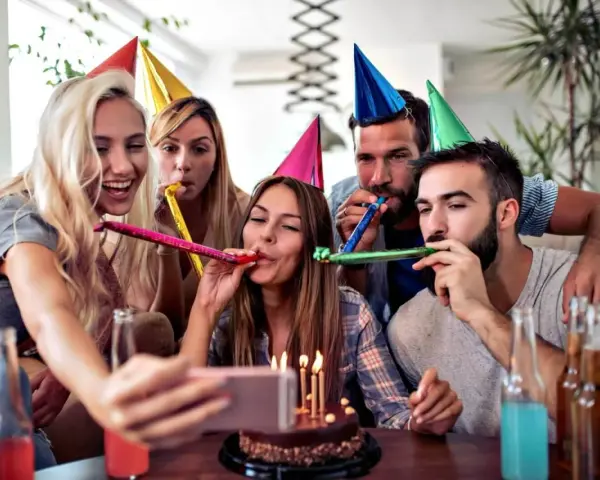 Radosne urodziny przyjaciółki! Grupa przyjaciół w kolorowych czapkach świętuje, dmucha w trąbki i robi selfie przy torcie.