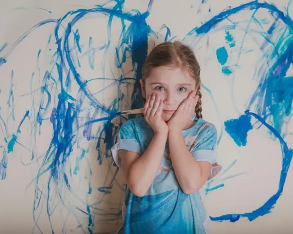 Dziewczynka z pędzlem w ręku, z dłońmi na policzkach, na tle niebieskich mazów. Czy asperger to choroba psychiczna?