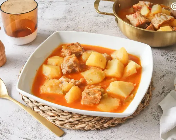 Un reconfortante plato de patatas con costillas en una salsa rojiza, servido en un cuenco blanco.