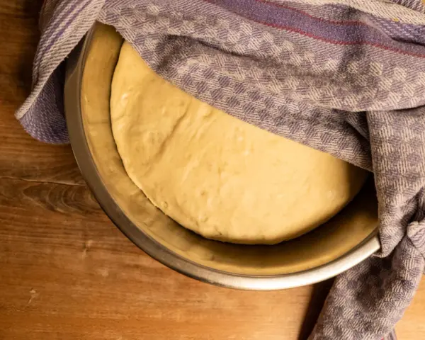 Ein geschmeidiger Teig ruht in einer Schüssel, bedeckt mit einem karierten Tuch, bereit für das Backen.