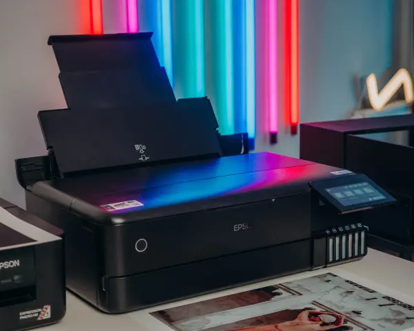 Czarna drukarka Epson z kolorowym ekranem, idealna do drukowania zdjęć. Obok stoi starszy model.