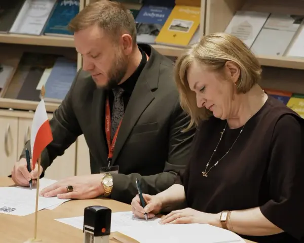 Para podpisująca dokumenty, z polską flagą obok. W tle półki z książkami, w tym te dotyczące slawistyki.