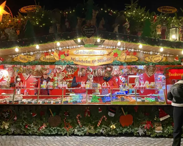 A vibrant Dresden Christmas market stall overflowing with gingerbread hearts, candied nuts, and colorful sweets. Festive lights twinkle above the tempting displays.
