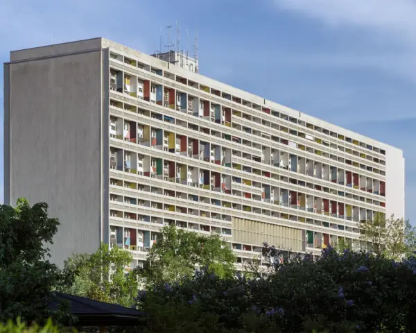 Długi budynek mieszkalny w stylu modernizm, z rzędami balkonów w kolorowych obramowaniach, otoczony zielenią.