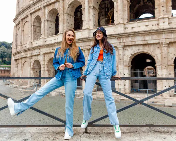 Dwie kobiety w jeans total look pozują przed Koloseum. Jedna ma rozpuszczone włosy i szeroki uśmiech, druga w kapeluszu bucket hat.