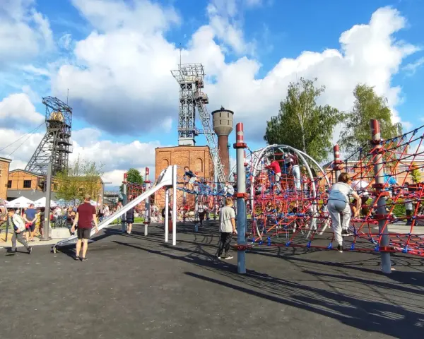 Rybnik atrakcje: plac zabaw dla dzieci z linowymi konstrukcjami i zjeżdżalnią, w tle wieże szybowe kopalni.