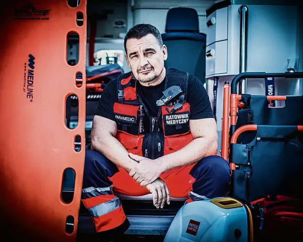 Ratownik medyczny w kamizelce z napisem "Paramedic" i "Ratownik Medyczny" siedzi w karetce obok noszy.