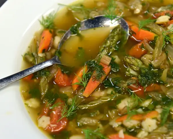 Un fumante minestrone di verdure, ricco di carote, fagioli e erbe aromatiche, servito in una ciotola bianca con un cucchiaio.