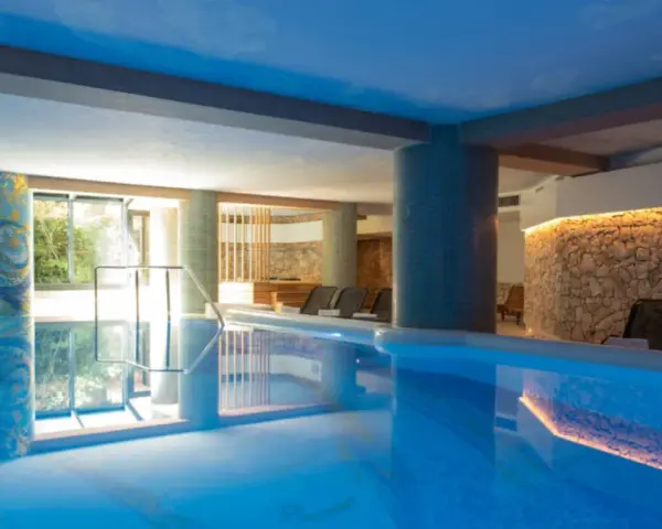 Indoor swimming pool with lounge chairs and a stone wall with warm lighting.