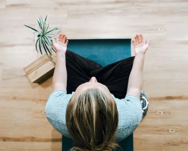 Person mediterar på en yogamatta, en grön växt och träblock bredvid. Perfekt för avkoppling på arbetsplatsen.