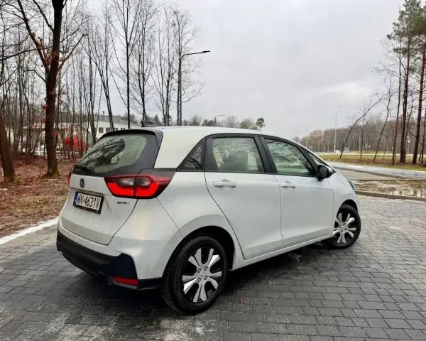 Biały Honda Fit stoi na mokrej, brukowanej nawierzchni. W tle drzewa i budynki.