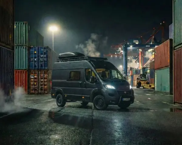 Mörk Fiat Ducato står vid en hamn, omgiven av containrar. Rök stiger från marken, kanske ett tecken på fiat ducato automatlåda problem.
