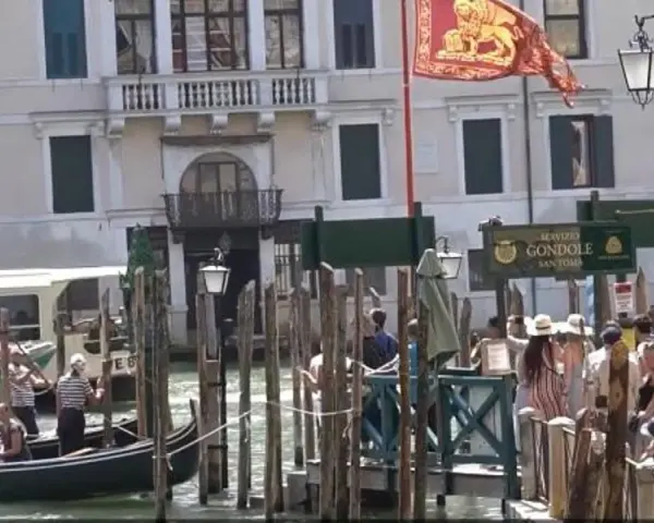 Un gruppo di innamorati a Venezia attende una gondola, con un vaporetto sullo sfondo e il leone di San Marco che sventola.