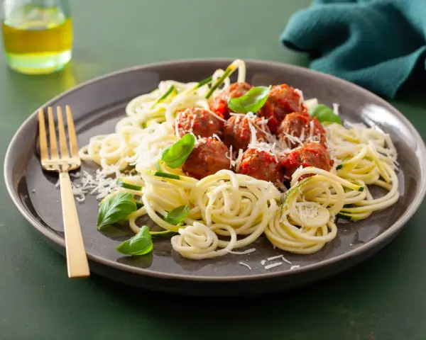 Pyszna keto kolacja: klopsiki w sosie pomidorowym na cukinii spaghetti, posypane parmezanem i listkami bazylii. Złoty widelec czeka.