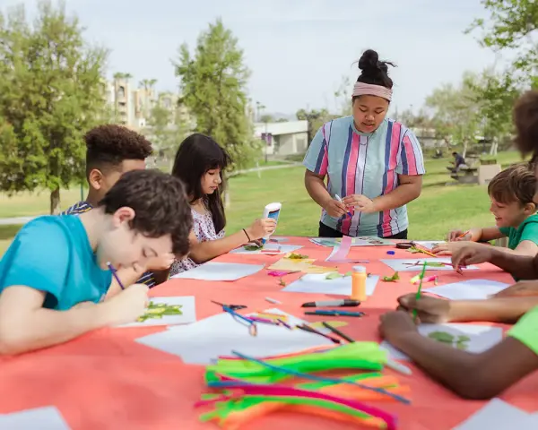 Des enfants créent des œuvres d'art en plein air autour d'une table recouverte d'une nappe orange.