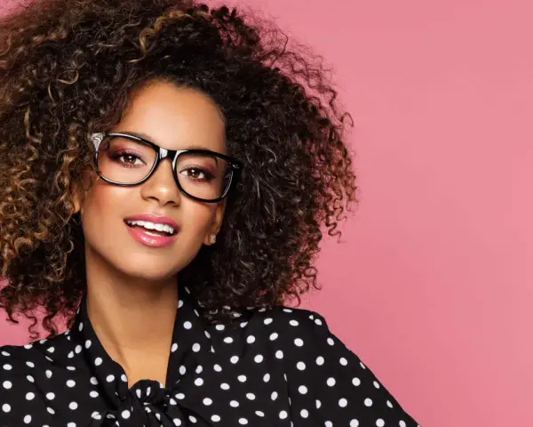 Une jeune femme aux cheveux bouclés porte un maquillage éclatant et des lunettes noires, souriant sur fond rose.