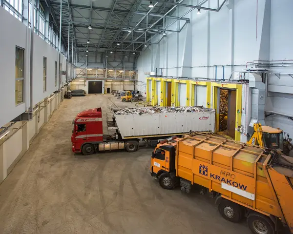 Wielka hala przemysłowa, gdzie trwa termiczne przekształcanie odpadów. Ciężarówki z odpadami czekają na rozładunek, a koparka przygotowuje materiał do przetworzenia.