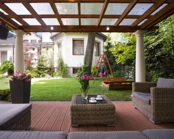 Drewniana pergola z przezroczystym dachem, tworząca zacienione miejsce do wypoczynku na tarasie. W tle ogród z huśtawką i piaskownicą.