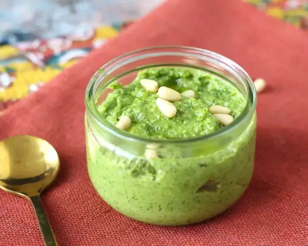 Frisches Rucola Pesto in einem Glas, garniert mit Pinienkernen. Daneben eine goldene Löffel.