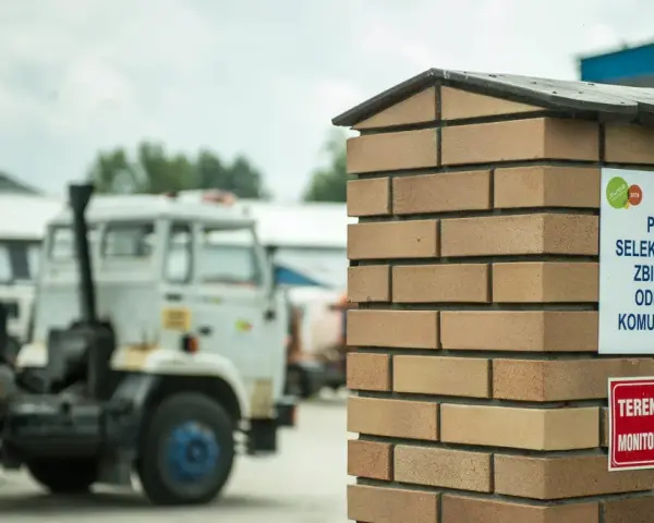 Tablica informuje, co to jest pszok – punkt selektywnego zbierania odpadów komunalnych. W tle widać ciężarówki.
