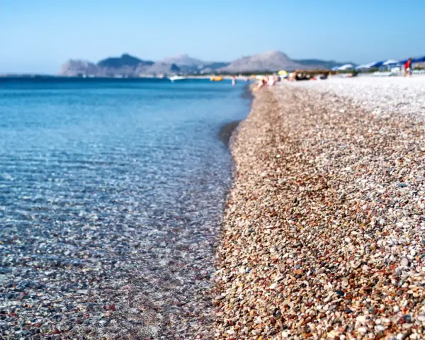 Rodos: turkusowe morze obmywa kamienistą plażę, w tle góry i plażowicze. Idealne miejsce na wakacje.