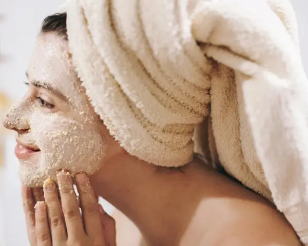 Kobieta z ręcznikiem na głowie nakłada na twarz peeling. To domowy sposób na gładką skórę.