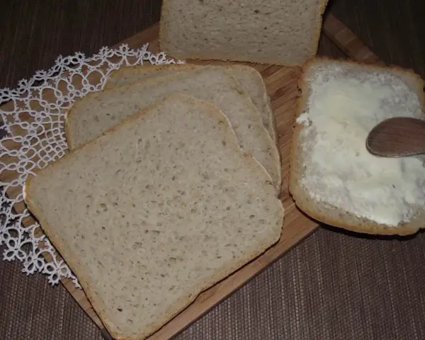 Chleb mieszany pokrojony w kromki, jedna z nich smarowana masłem drewnianą łyżką.