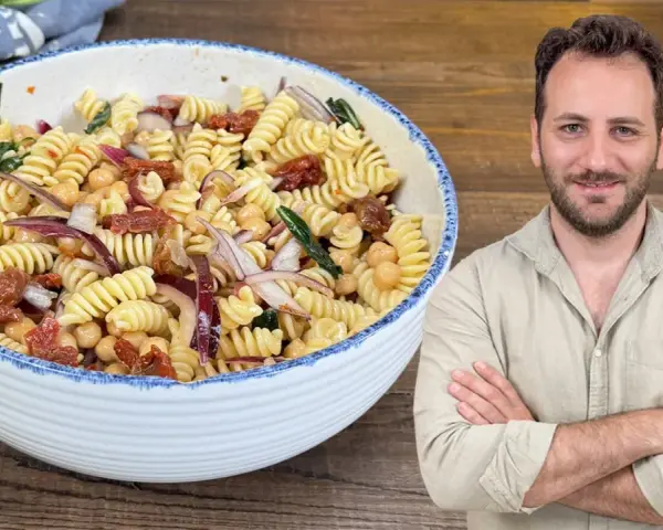 Un uomo sorride accanto a una ciotola di pasta fredda con ceci, pomodori secchi e cipolla rossa.