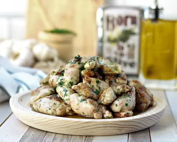 Deliciosas mollejas de cordero salteadas con perejil fresco, servidas en un plato de madera. Al fondo, aceite de oliva y ajos.