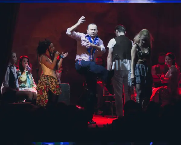 Scena teatralna, gdzie mężczyzna tańczy, a kobiety śpiewają. Atmosfera jak po pożarze w burdelu, pełna emocji i dramatyzmu.