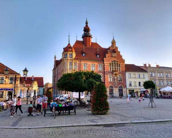 Rynek w Tarnowskich Górach tętni życiem. Ludzie spacerują, siedzą na ławkach, a dzieci bawią się na bruku. W tle widać zabytkowy ratusz.