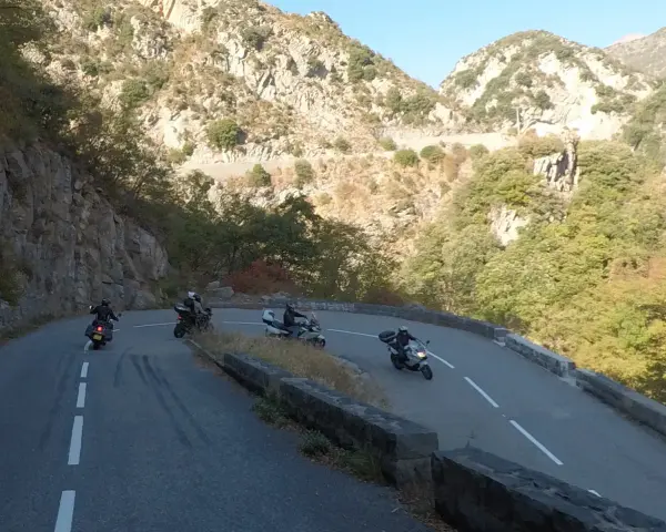 Motociclisti affrontano le curve del Col de Turini, immersi in un paesaggio montano roccioso e verdeggiante.