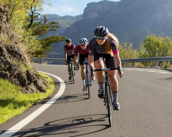Three cyclists race up a mountain road. The lead cyclist, a woman, is what is a sportive person, focused and determined.