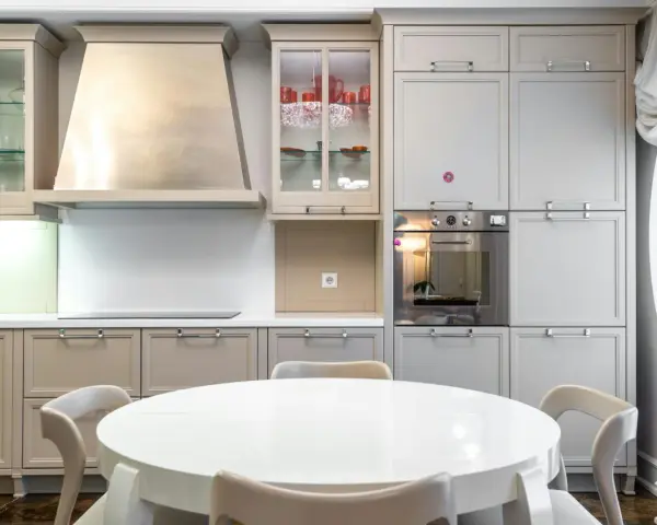 Cocina moderna con mesa redonda blanca y sillas. Gabinetes grises y beige, campana extractora de acero inoxidable. Ideal para ver catálogos de cocinas y precios.