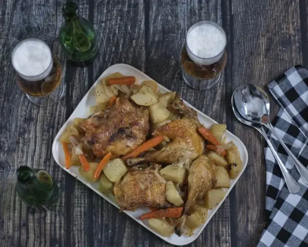 Pollo asado con patatas y zanahorias en una bandeja blanca, con cerveza y servilleta a cuadros.