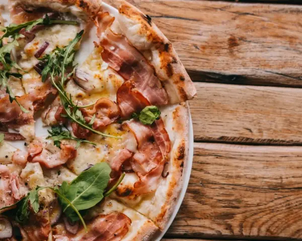 Pyszna pizza carbonara z chrupiącym boczkiem, rukolą i cebulą na drewnianym stole.
