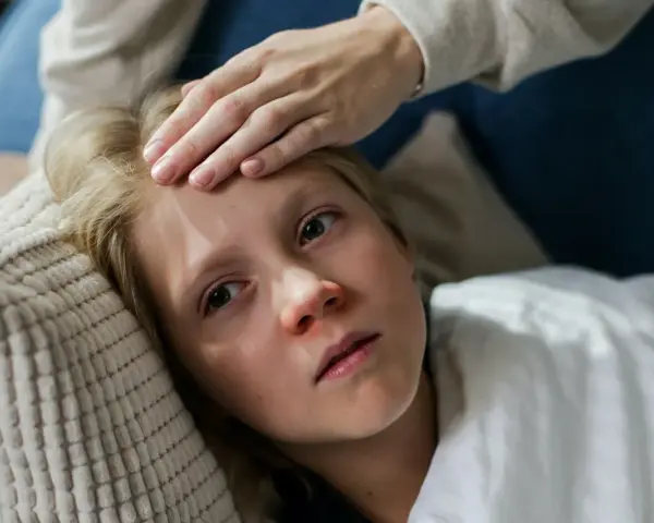 Dziecko z gorączką leży na poduszce, a ręka mamy sprawdza mu czoło. Czy dziecko z gorączką może wychodzić na dwór?