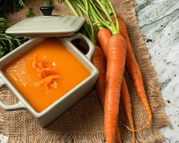 Un tazón de cremoso puré de zanahoria, adornado con hebras de zanahoria rallada, junto a zanahorias frescas y verdes.