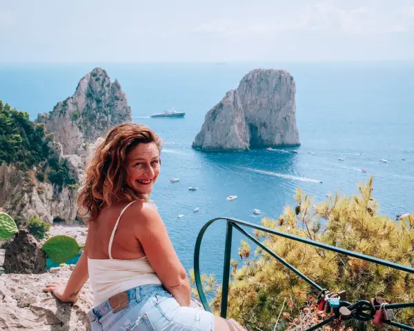 Donna sorridente con vista sui Faraglioni, un'icona dell'isola di Capri. Il mare azzurro e le barche completano il paesaggio.