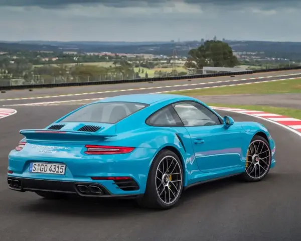Błękitne Porsche 911 Turbo S na torze wyścigowym, z widokiem na miasto w tle.