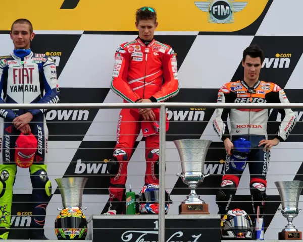 Podio con tre motociclisti famosi in tuta: Rossi, Stoner e Lorenzo. Vittoria celebrata con trofei.